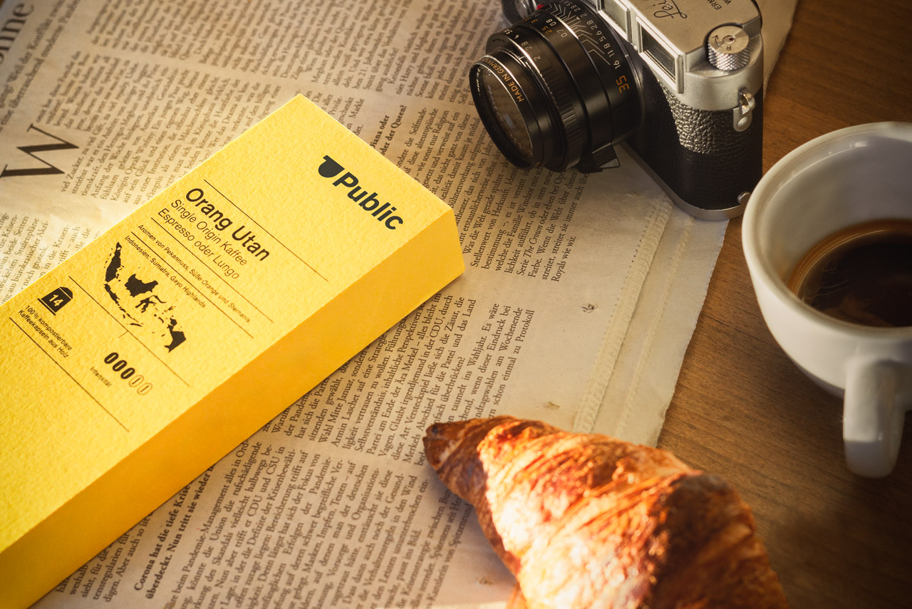 Eine Packung Kaffeekapseln liegt auf einer Zeitung. Daneben eine Leica Kamera und ein Croissant.